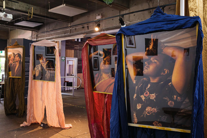 Several large photographic portraits hang from the ceiling of the gallery draped in coloured cloth.