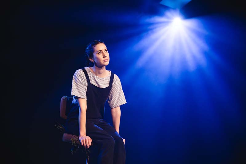 A woman sits alone on a darkened stage under a spotlight. She is wearing a stripped t-shirt and black overalls.