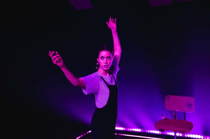 A woman waves her arms in the air, alone on a darkened stage under a spotlight. She is wearing a stripped t-shirt and black overalls.