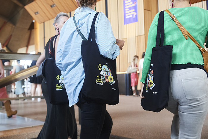 Close up of three women holding All About Women tote bags. The bags are black with bright graphics.