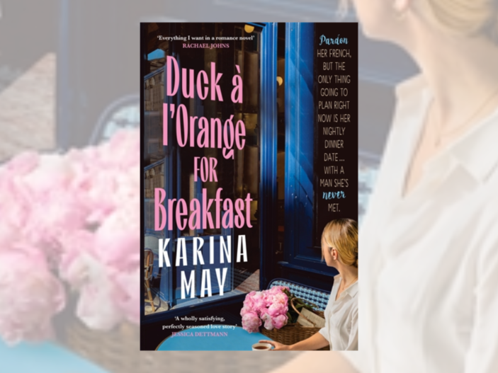 The cover of Duck a' l'Orange for Breakfast by Karina May has the title in a light pink and the author's name in white capital letters. Next to these words there is a blonde woman sitting at a table with a cup of coffee and a basket of pink flowers looking out the window deep in thought