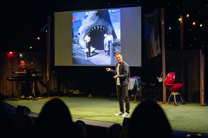 Rove McManus stands on a stage in front of a slide of a giant fake shark.