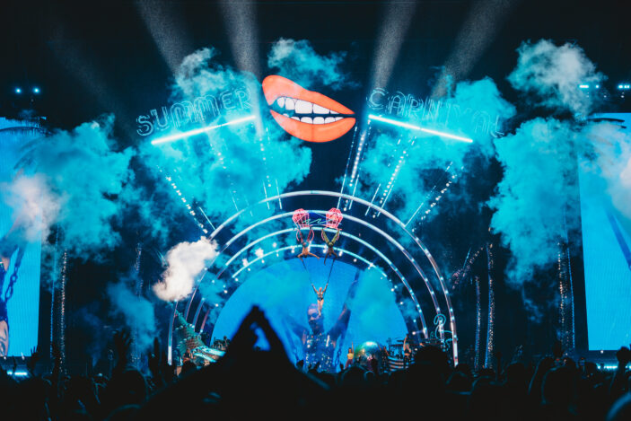 Main stage of Pink's Summer Carnival stage with Pink in a harness with two men above her in harnesses and blue smoke